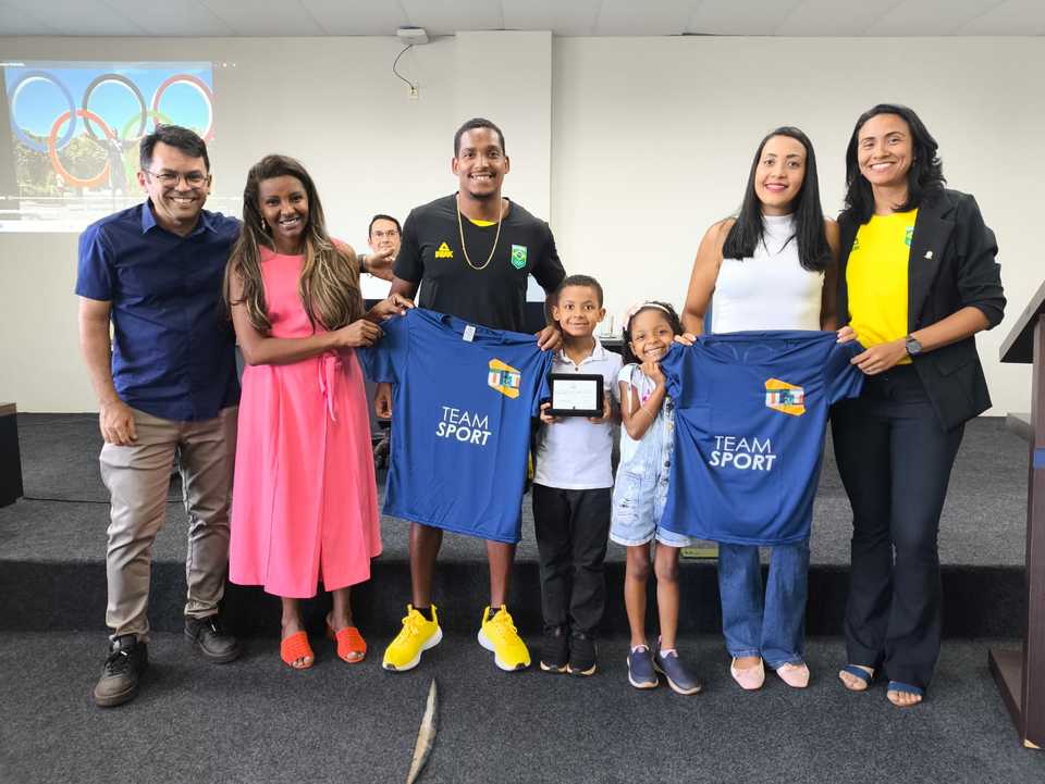 Foto colorida com representantes da Proex, Jack, sua esposa e filhos segurando a placa e camisetas da universidade. 